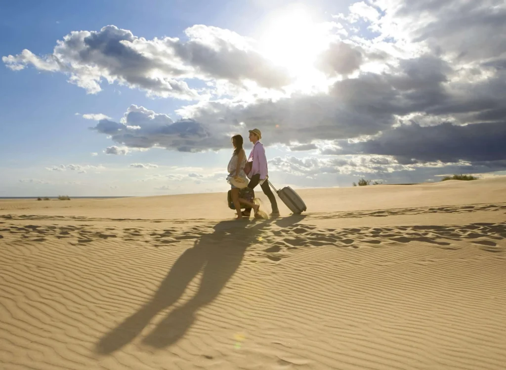 Traveling couple walking on a sandy beach while dragging luggage after travel health & immunization consultation